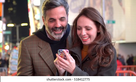 Young Happy Couple Viewing Smart Cell Phone In Times Square New York At Night. Share Memories And Pictures On Social Media With Online Mobile  App