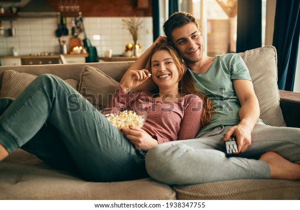 Young happy couple watching movie on\
TV and eating popcorn while relaxing in the living room.\
