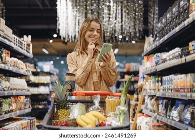 Young happy customer woman wear casual clothes use mobile cell phone shopping at supermaket store grocery shop buy with trolley cart choose products in hypermarket. Purchasing food gastronomy concept - Powered by Shutterstock