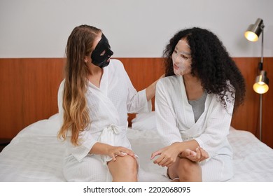 Young Happy Diversity Ethnic Beautiful Woman In Bathrobe Sit On Bed And Doing Cosmetic Black And White Facial Mask For Skin Care And Treatment. Women Lesbian Couple Daily LGBTQ Lifestyle Concept.