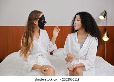 Young Happy Diversity Ethnic Beautiful Woman In Bathrobe Sit On Bed And Doing Cosmetic Black And White Facial Mask For Skin Care And Treatment. Women Lesbian Couple Daily LGBTQ Lifestyle Concept.