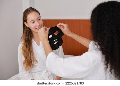 Young happy diversity ethnic beautiful woman in bathrobe sit on bed and doing cosmetic black facial mask for skin care and treatment. Women  couple daily - Powered by Shutterstock