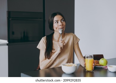 Young happy dreamful minded housewife woman 20s wearing casual clothes beige shirt eat oatmeal porridge muesli in morning cooking food in light kitchen at home alone. Healthy diet lifestyle concept. - Shutterstock ID 2099292835