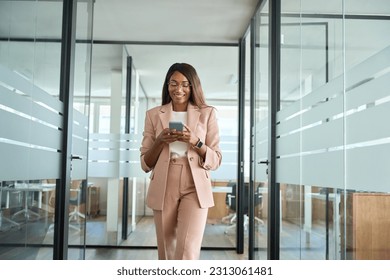 Young happy elegant busy professional African American business woman company executive manager worker wearing suit using cell phone walking in office hall holding mobile phone and working.