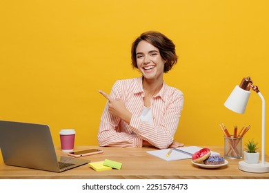 Young happy employee business woman wear casual shirt sit work at office desk with pc laptop point fingers aside on area mockup isolated on plain yellow color background. Achievement career concept