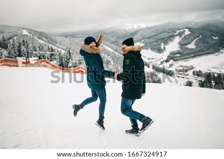young happy european couple in dark jackets and hats jumping, running in hats in winter in mountains