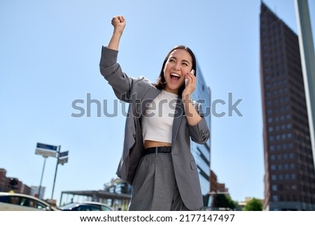 Young happy excited Asian business woman entrepreneur winner standing on city street talking on phone celebrating work success, succeed in career and financial goals raising hand in yes gesture.