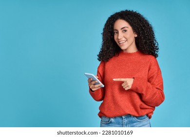 Young happy excited latin woman pointing at mobile phone isolated on blue background. Smiling female model holding cellphone using cell presenting advertising new trendy application concept. - Shutterstock ID 2281010169