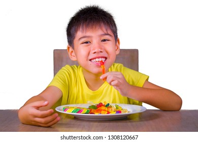 Young Happy And Excited Male Kid Smiling Cheerful Eating Dish Full Of Candy And Lollipop Sitting At Table Isolated On White Background In Sugar Addiction And Abuse And Sweet Unhealthy Nutrition 