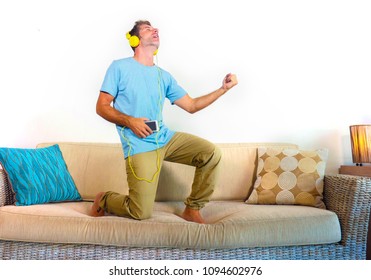 Young Happy And Excited Man Jumping On Sofa Couch Listening To Music With Mobile Phone And Headphones Playing Air Guitar Crazy Having Fun At Home Living Room In Audio Fan Lifestyle Concept