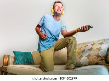 Young Happy And Excited Man Jumping On Sofa Couch Listening To Music With Mobile Phone And Headphones Playing Air Guitar Crazy Having Fun At Home Living Room In Audio Fan Lifestyle Concept