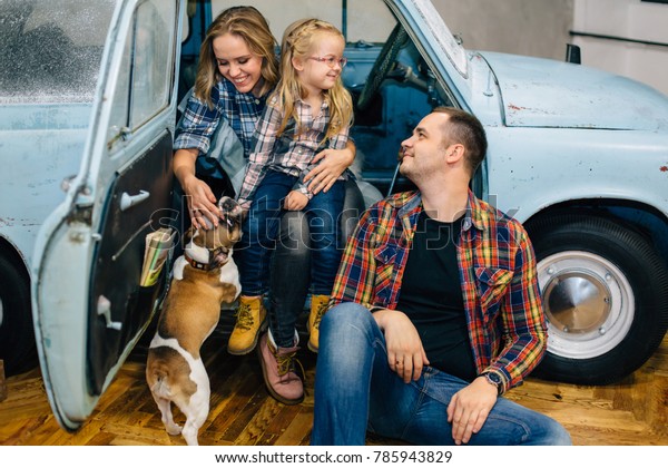 Young happy family in checked shirts sittng near\
blue retro car with dog. Couple in love with cute daughter having\
fun with french bulldog