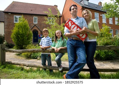 Young Happy Family Holding Sold Sign Outside Their New House.