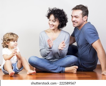 Young Happy Family With The Mother, Father And A Funny Daughter Sitting On A Wood Floor Playing Clapping