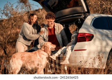 A young happy family on a picnic with their beloved little daughter and dog sitting in the back of a car on a blanket on a weekend having fun together. - Powered by Shutterstock