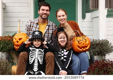 Young happy family parents with kids in Halloween costumes holding carved pumpkins jack o lanterns and smiling at camera while sitting on house porch and waiting to trick or treat