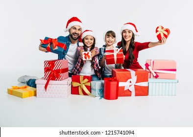 Young Happy Family In Santa Hats With Pile Of Christmas Presents Show It Into The Camera Isolated On White Background. New Year Holidays