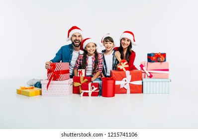 Young Happy Family In Santa Hats With Pile Of Christmas Presents Show It Into The Camera Isolated On White Background. New Year Holidays