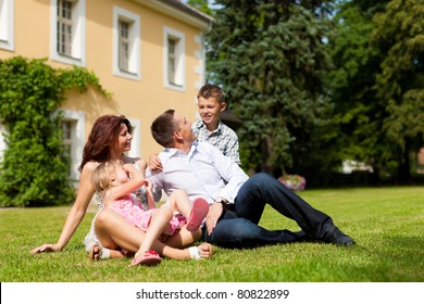 Young Happy Family Sitting In The Sun On The Lawn In Front Of Their New Home - A Villa