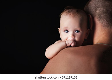 Young Happy Family In The Studio On A Dark Background In Dark Clothes Holding A Baby On The Hands And Looks At Him.