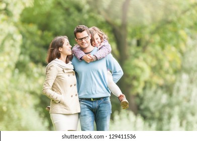 Young Happy Family Of Three Having Fun Together Outdoor. Pretty Little Daughter On Her Father Back. Parents And Girl Look Happy And Smile. Happiness And Harmony In Family Life. Family Fun Outside.