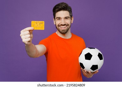Young happy fan smiling man he wear orange t-shirt cheer up support football sport team hold in hand soccer ball mock up of credit bank card watch tv live stream isolated on plain purple background - Shutterstock ID 2195824809
