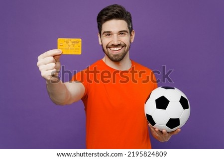 Young happy fan smiling man he wear orange t-shirt cheer up support football sport team hold in hand soccer ball mock up of credit bank card watch tv live stream isolated on plain purple background