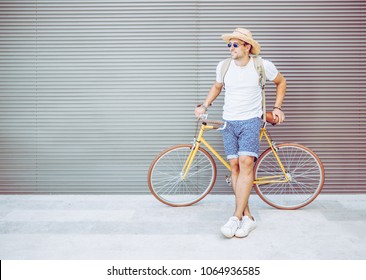 Young Happy Fashion Hipster Man With Cool Yellow Vintage Bicycle. Male Leaning At His Bike While Standing Against Grey Background. Outdoors Portrait Of Handsome Guy In Summer Clothes.     