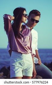 Young Happy Fashionable Couple Resting By The Sea On Sunset. The Man And Woman Wearing In Romantic Sexy Summer Clothes And  In Stylish Trendy Sunglasses