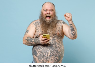 Young Happy Fat Pudge Obese Chubby Overweight Blue-eyed Bearded Man 30s Has Big Belly With Naked Tattooed Torso Point Finger On Mobile Cell Phone Do Winner Gesture Isolated On Pastel Blue Background.