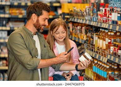 Young happy father with a child buying groceries in a supermarket using a smartphone, scanning a QR code. A man carries his daughter in a grocery cart, choosing goods and having fun. - Powered by Shutterstock