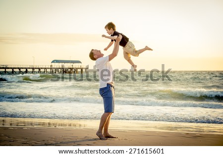 young happy father holding up in his arms little son putting him up at the beach in barefoot standing in front of sea waves wet sand having fun with the kid in Summer sunset coast
