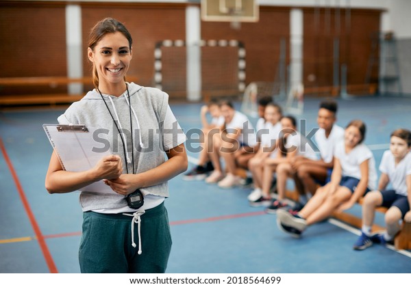 Young happy female\
coach looking at camera while having PE class with elementary\
students at school gym. 