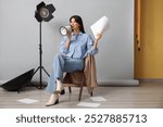 Young happy female film director with megaphone, script and studio lighting sitting on chair against grey background