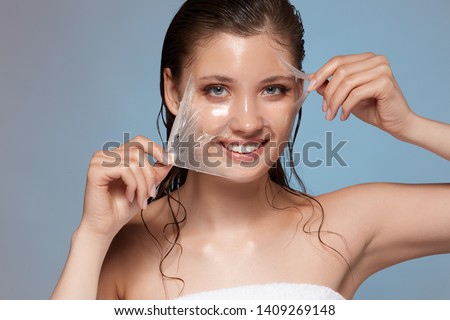 young and happy female peeling off facial mask and smiling to the camera, woman treats her face in bath towel