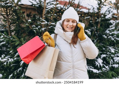 Young happy female in winter clothes with shopping bags standing in Christmas market with illuminated tree behind and speaking on mobile