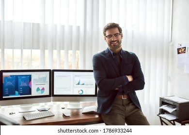 Young happy financial analyst in classic wear   eyeglasses leaning against table   smiling at camera while working and statistical data  graphs   charts computer in the modern office