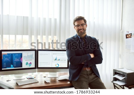Young happy financial analyst in classic wear and eyeglasses leaning against a table and smiling at camera while working with statistical data, graphs and charts on computer in the modern office