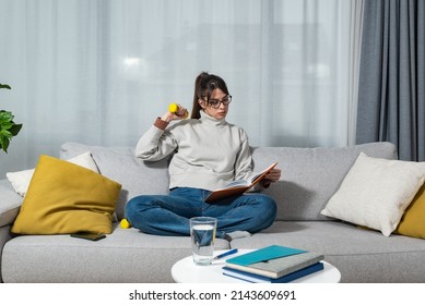 Young Happy Fitness College Student Woman Sitting At Home Study Reading Books To Educate Herself Holding Small Weight And Exercise With One Hand. Health And Motivated Life Concept