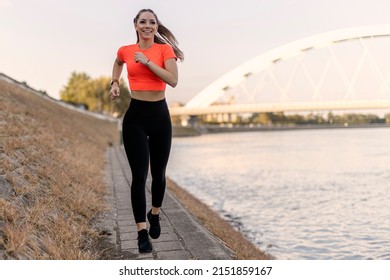 Young happy focused fitness girl in black yoga pants and orange short shirt jogs on riverbank during the day. Front view.