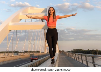 Young Happy Focused Fitness Girl In Black Yoga Pants And Orange Short Shirt Practice Balance On Bridge Fence During The Day. Front View.