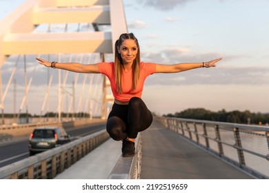 Young Happy Focused Fitness Girl In Black Yoga Pants And Orange Short Shirt Practice Balance On Bridge Fence During The Day. Front View.