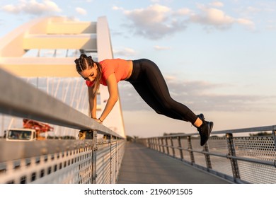 Young Happy Focused Fitness Girl In Black Yoga Pants And Orange Short Shirt Jumps From Bridge Fence On Footpath. Front View.