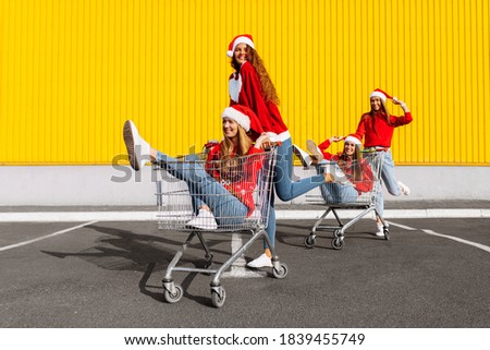 Young happy friends in Christmas sweaters and Santa Claus hats are having fun on a shopping trolley, against the background of a yellow wall of a shopping center, shopping concept, Christmas, New Year