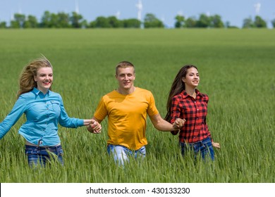 Young Happy Friends Running On Green Wheat Field. One Guy And Two Girls Are Jumping, Having Fun.