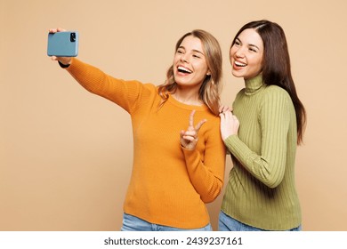 Young happy friends two women they wear orange green shirt casual clothes together doing selfie shot on mobile cell phone post photo on social network show v-sign isolated on plain beige background