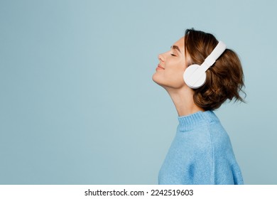 Young happy fun caucasian woman wear knitted sweater headphones listen to music with closed eyes have fun isolated on plain pastel light blue cyan background studio portrait. People lifestyle concept