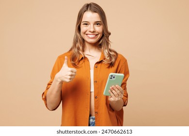 Young happy fun Caucasian woman she wear orange shirt casual clothes hold in hand use mobile cell phone show thumb up isolated on plain pastel light beige background studio portrait. Lifestyle concept - Powered by Shutterstock