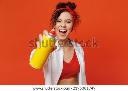 Young happy fun cool woman of African American ethnicity wear white shirt top hold paint spray bottle make graffiti stretch hand to camera isolated on plain orange background People lifestyle concept