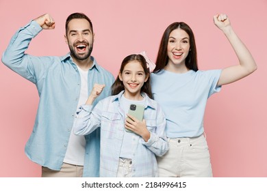 Young Happy Fun Parents Mom Dad With Child Kid Daughter Teen Girl In Blue Clothes Hold Use Mobile Cell Phone Do Winner Gesture Isolated On Plain Pastel Pink Background. Family Day Parenthood Concept